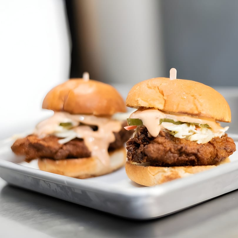 Two crispy chicken sliders with coleslaw, pickles, and sauce on a white tray.