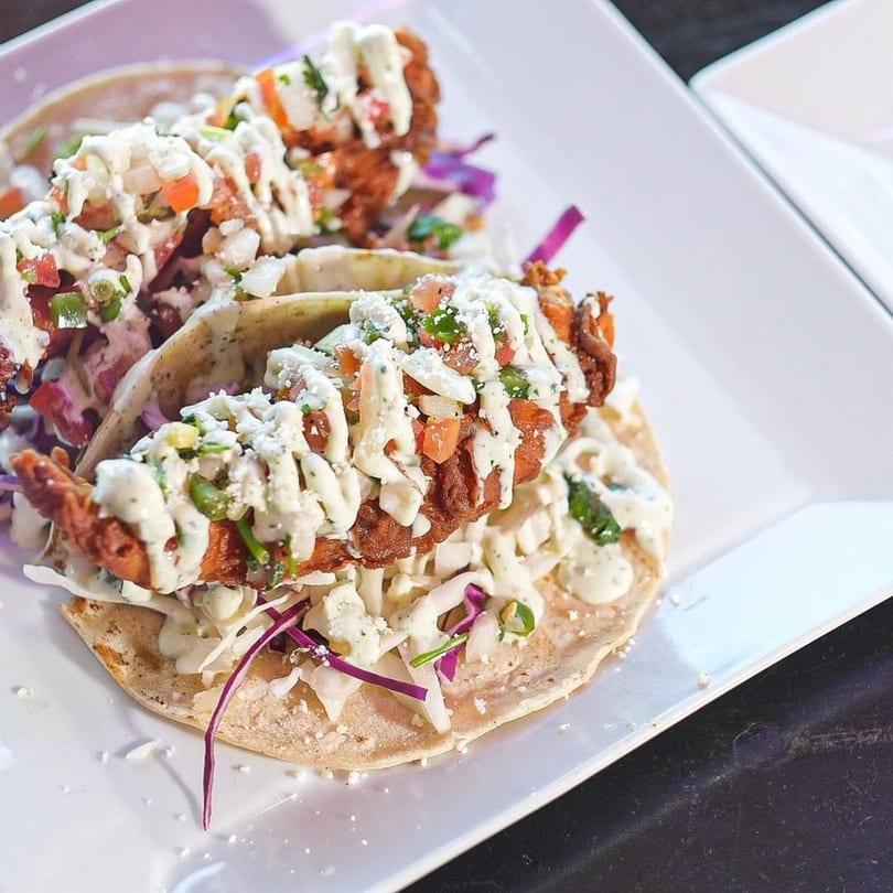 Two crispy fish tacos with white sauce, pico de gallo, and purple cabbage on a white plate.