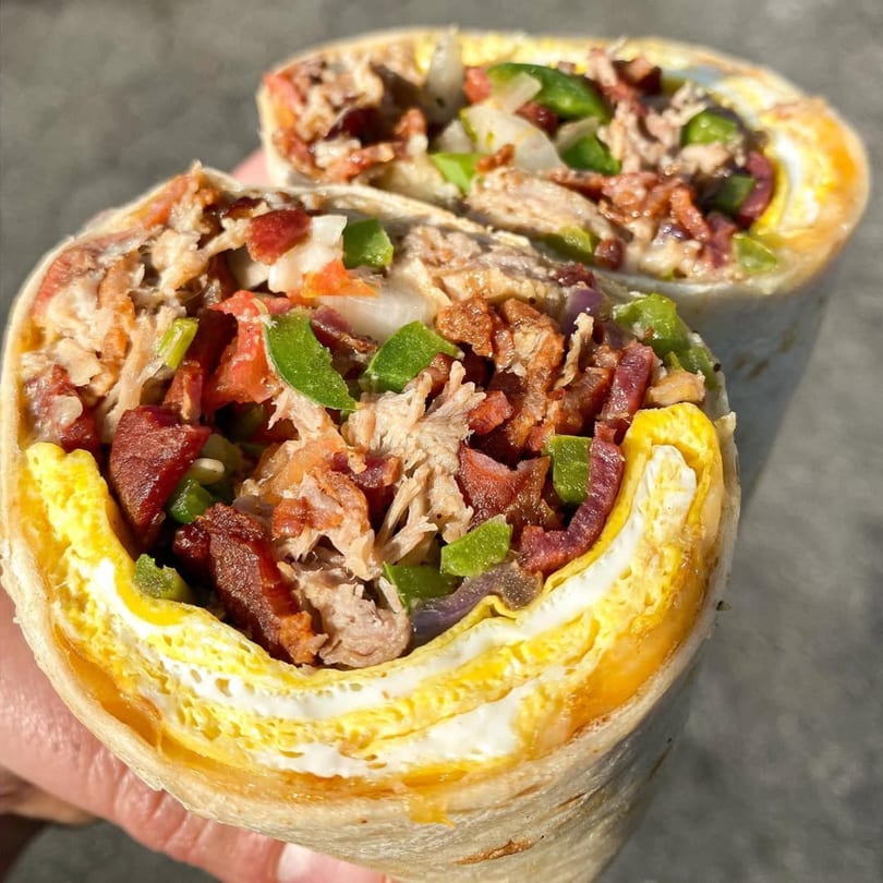 Two halves of a breakfast burrito filled with scrambled eggs, bacon, pulled pork, and green peppers.
