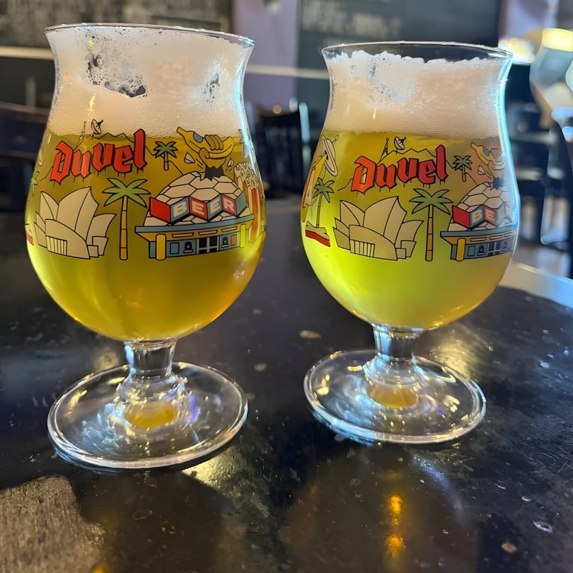 Two Duvel beer glasses filled with golden beer and foamy heads on a dark table.